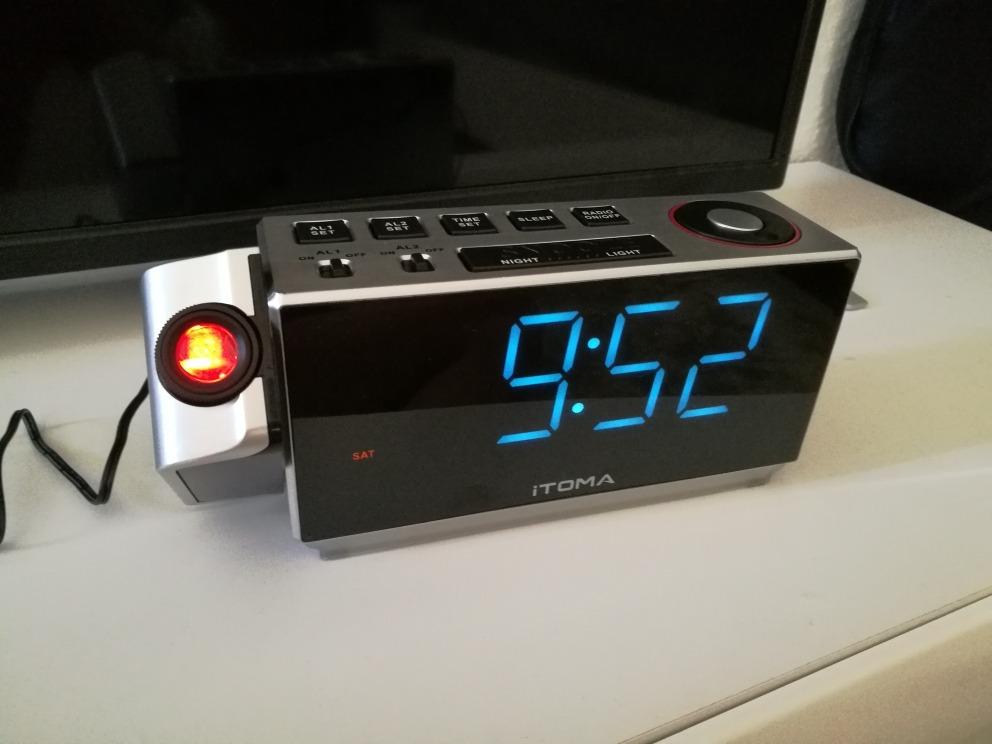 Radio réveil posé sur une surface blanche, le bras du projecteur est sorti, l'écran affiche 9h52.