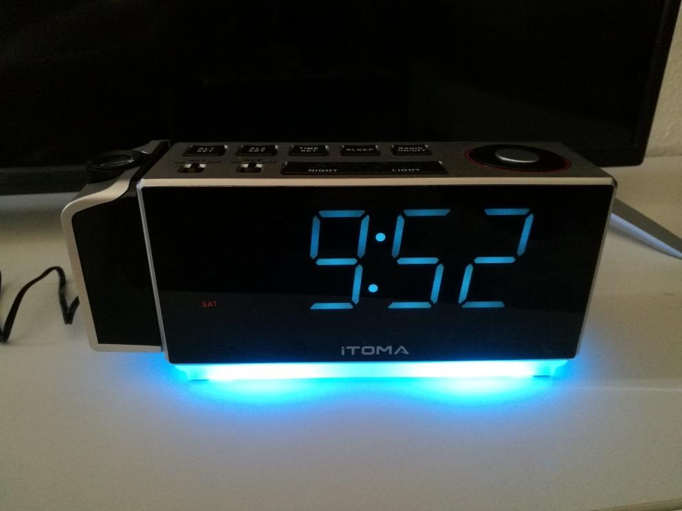 Radio réveil posé sur une surface blanche, le socle est allumé en bleu, l'écran affiche 9h52.