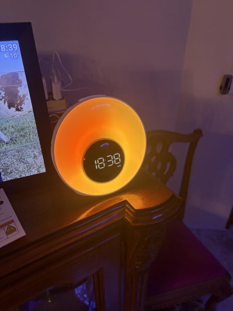 Le réveil lumière en situation dans un intérieur sombre avec un éclairage orangé, posé sur une commode en bois. On voit aussi...