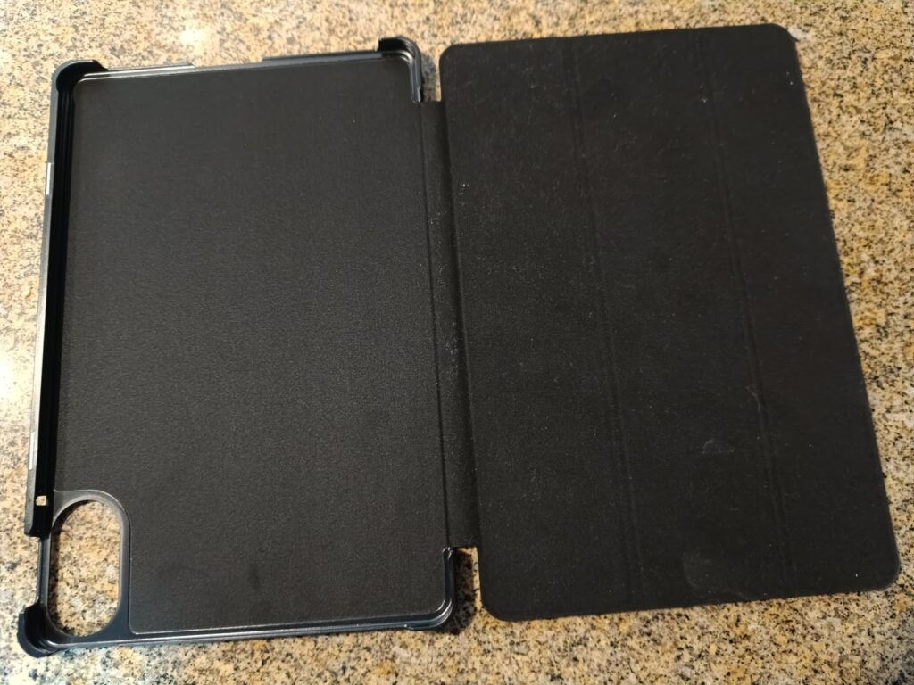 La housse de protection de tablette, ouverte, posée à plat sur une surface marron clair. On voit l'intérieur de la coque et l...