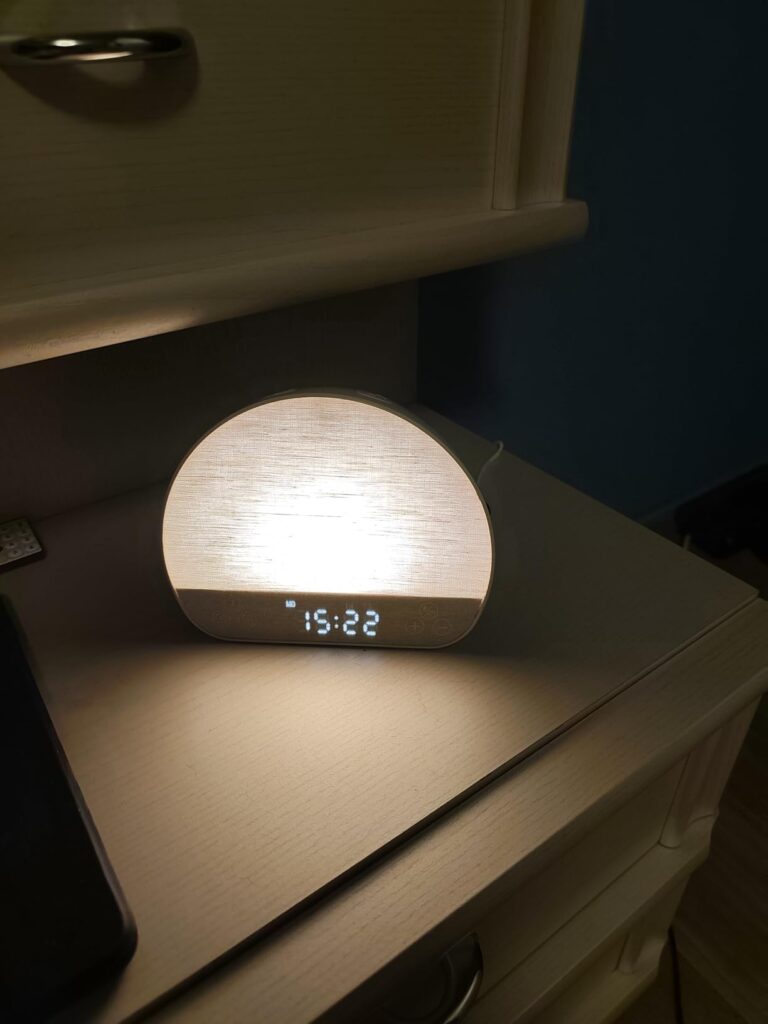 Le réveil-lumière est posé sur une table de chevet, allumé, avec l'heure affichée, la lumière est blanche.