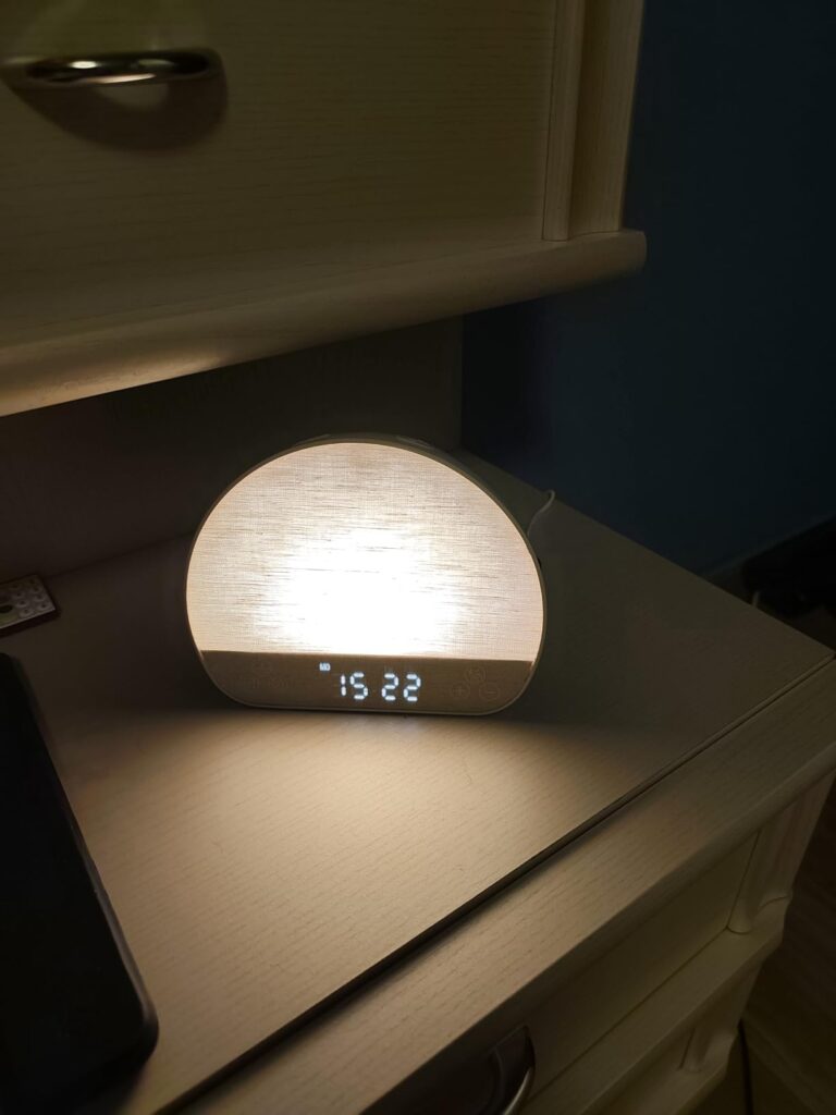 Le réveil-lumière est posé sur une table de chevet, allumé, avec l'heure affichée, la lumière est blanche.