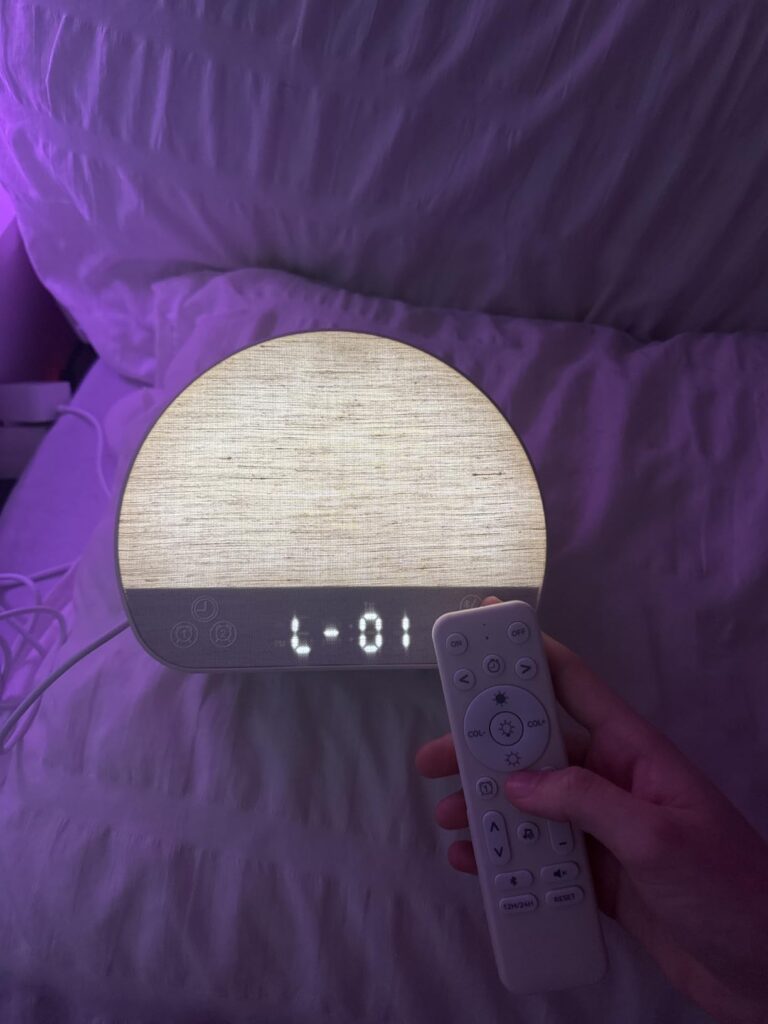 Le réveil-lumière est posé sur un lit avec une personne qui tient la télécommande pour le contrôler, la lumière du réveil est...