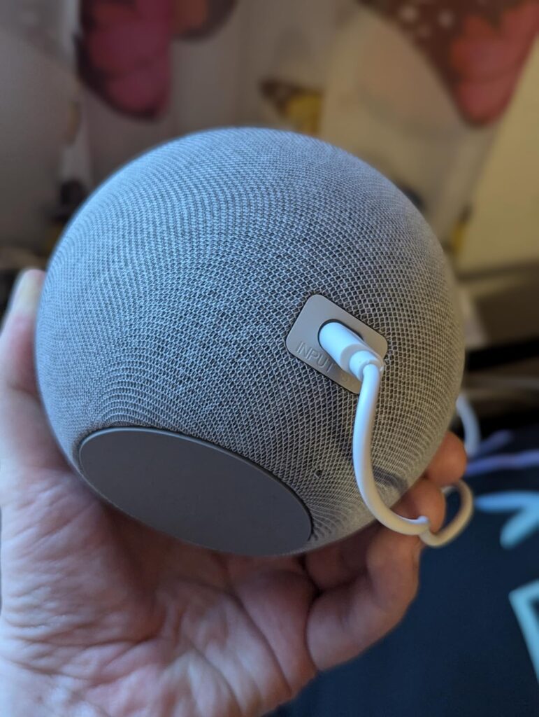 L'enceinte est tenue dans une main, le câble de chargement est branché à l'arrière.