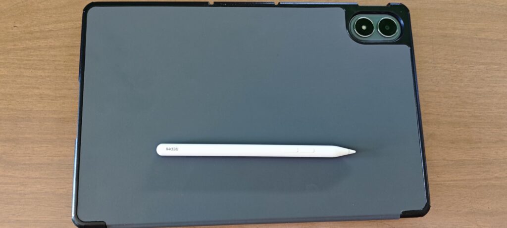 Vue de dos de la tablette dans son étui, avec un stylet posé dessus. On voit l'objectif photo en haut à droite.