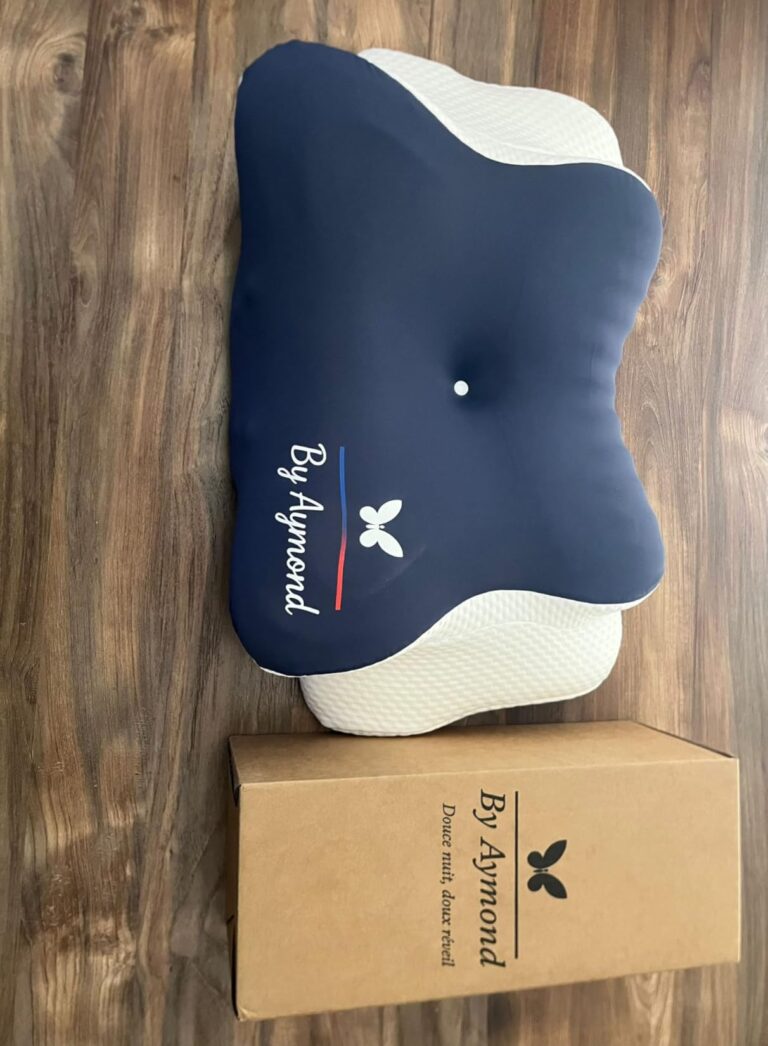 Le coussin By Aymond en forme de papillon, de couleur bleu marine et blanc, posé sur un plancher en bois, avec sa boîte d'emb...