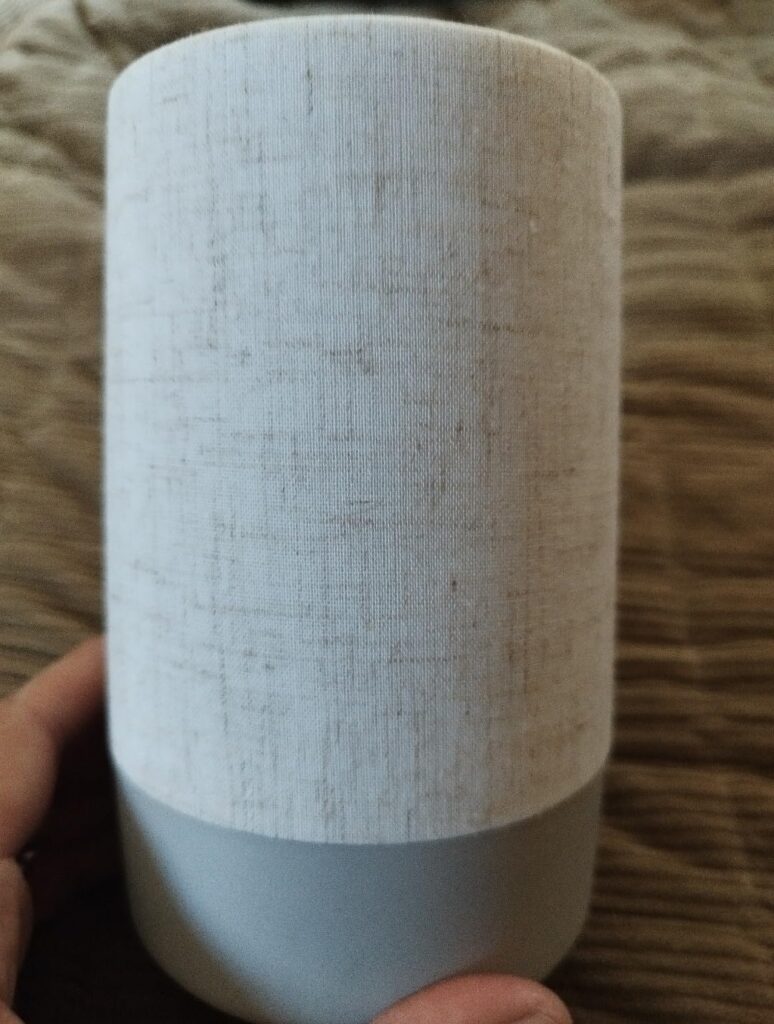 Vue générale de l'appareil en main, en forme de cylindre avec un revêtement en tissu gris clair et une base gris foncé.
