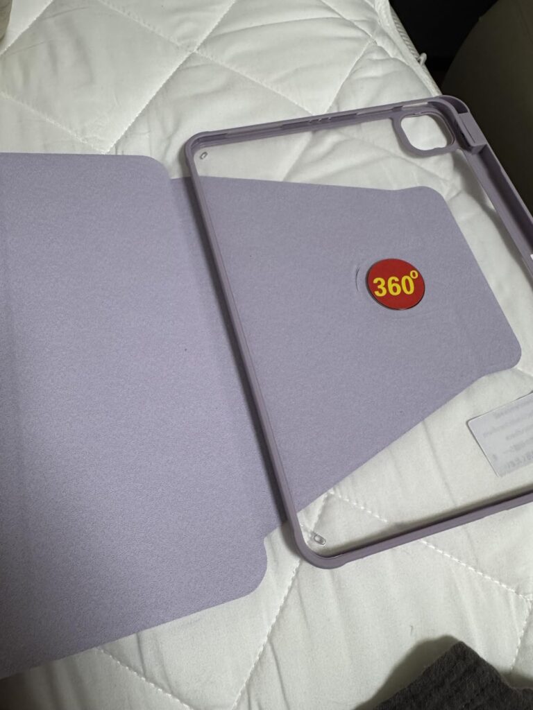 La coque de protection est ouverte sur un fond clair (un lit avec un drap blanc). La coque est de couleur mauve/violette et l...