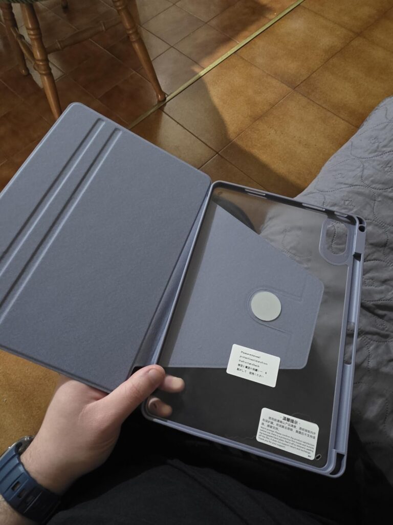 Une main tient la coque de protection ouverte sur un fond sombre (canapé ou plaid). La coque est de couleur mauve/violette et...