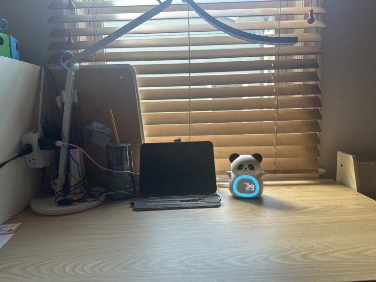 Le réveil panda est posé sur un bureau en bois, près d'une tablette et d'une lampe, devant une fenêtre avec des stores. Le ré...