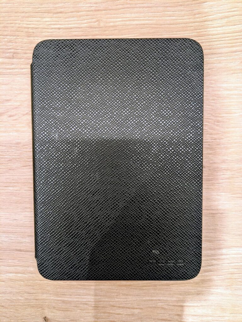 Vue de dessus d'une liseuse Kindle Paperwhite, recouverte d'un étui noir fermé. On voit un motif qui ressemble à du carbone.
