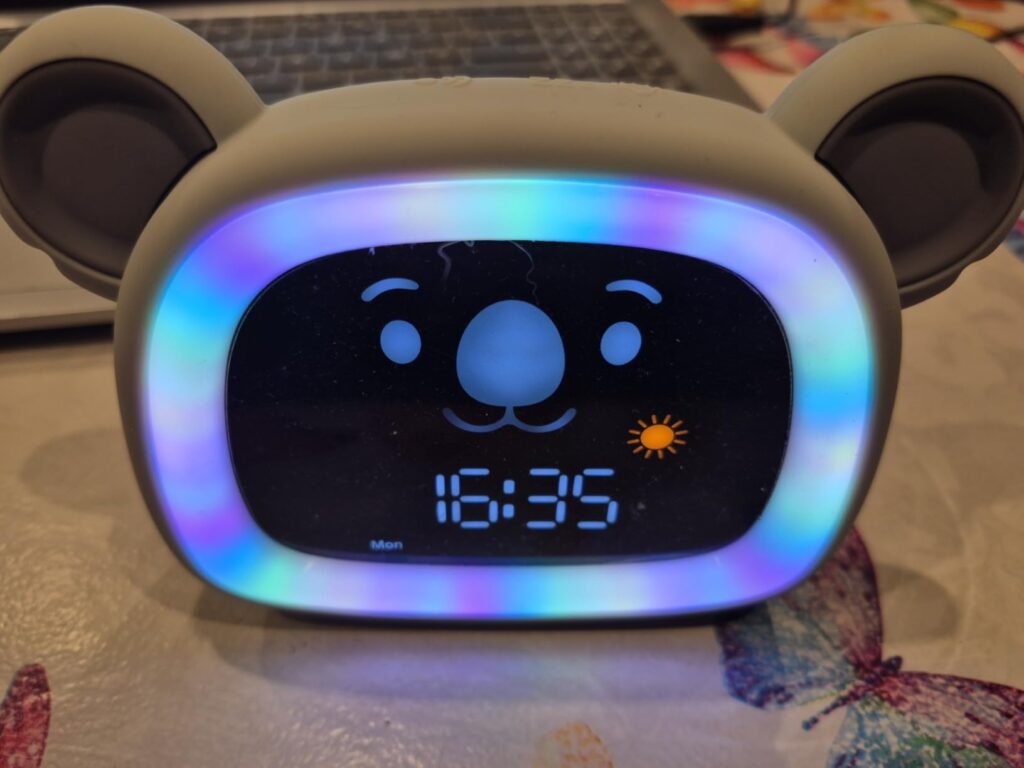 Gros plan sur l'écran du réveil-veilleuse, affichant 16:35 et un koala souriant. L'écran est entouré d'une lumière multicolor...