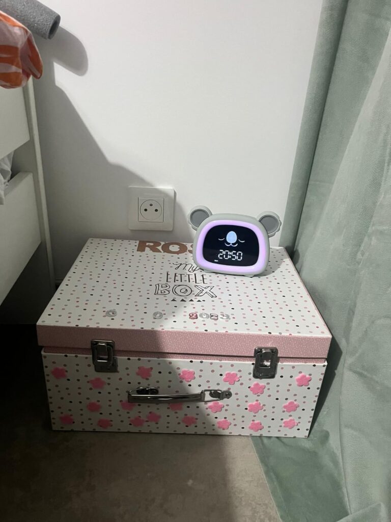 Le réveil-veilleuse est posé sur une petite malle de rangement, près d'une prise électrique, dans ce qui semble être une cham...