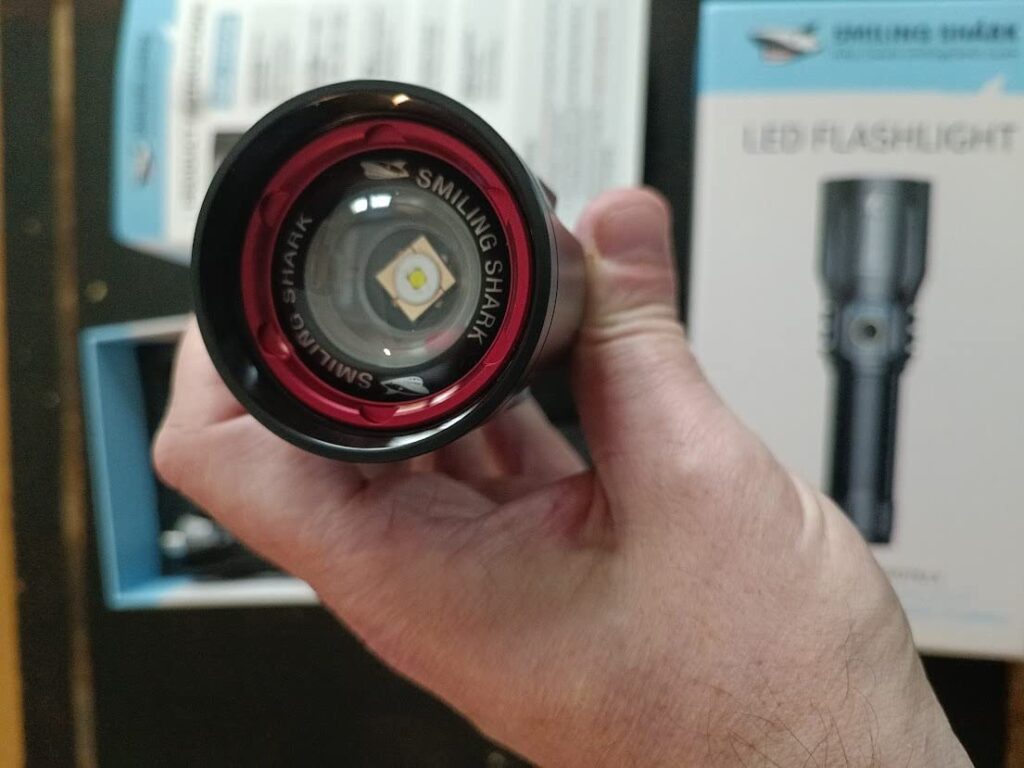 Vue de l'extrémité de la lampe torche, avec l'ampoule LED visible. Le texte 'SMILING SHARK' est lisible sur l'anneau rouge.