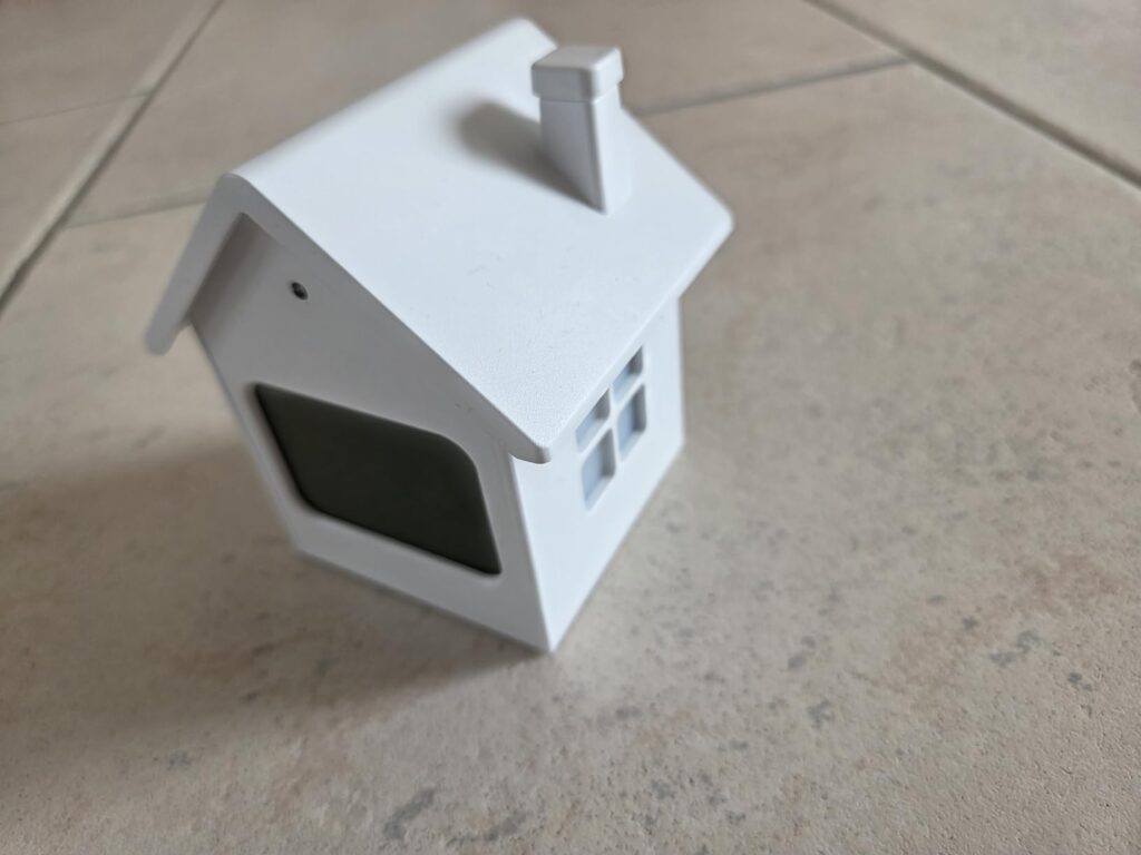 Vue de face et du côté droit du réveil en forme de maison, de trois quarts, posé sur un carrelage. L'écran est éteint.