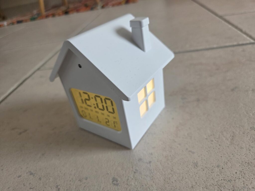 Vue de face et du côté droit du réveil en forme de maison, de trois quarts, posé sur un carrelage. La fenêtre latérale est al...