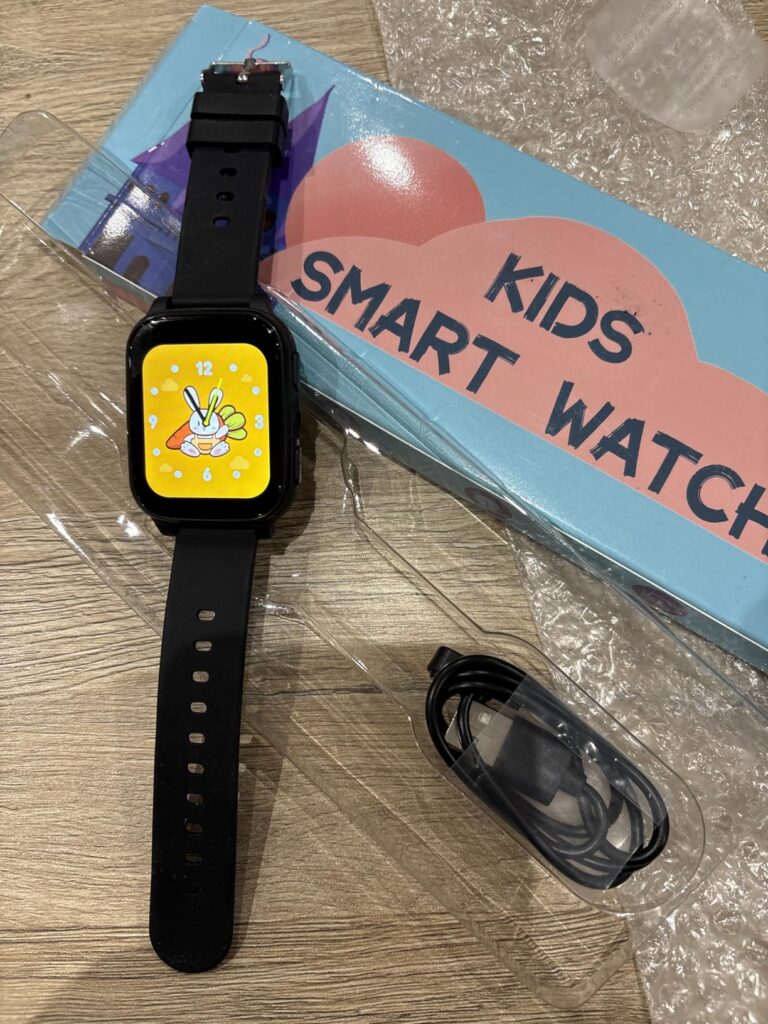 Montre enfant noire allumée et son câble de chargement, à côté de sa boîte, le tout sur un sol en bois