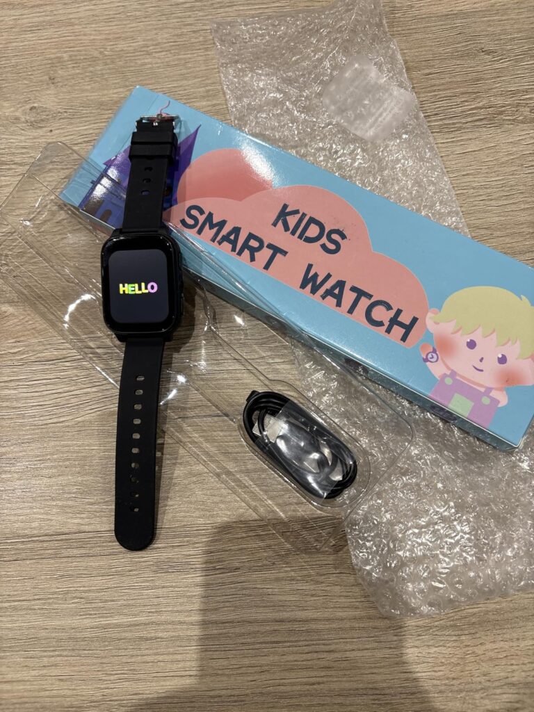 Montre enfant noire allumée et son câble de chargement, à côté de sa boîte, le tout sur un sol en bois