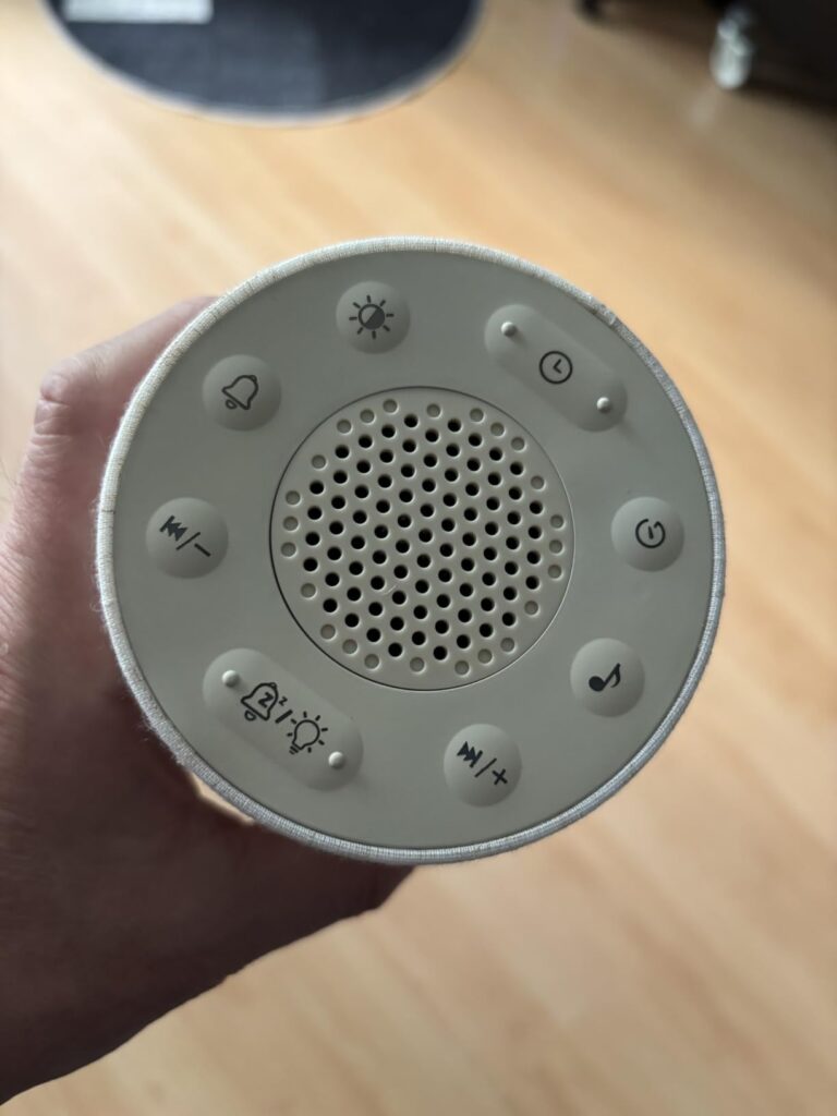 Vue du dessus du réveil-enceinte, montrant les boutons de contrôle et le haut-parleur. Une main tient l'appareil.