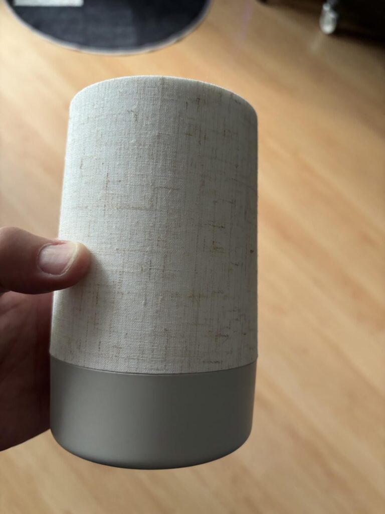 Vue générale du réveil-enceinte, avec un revêtement en tissu clair et une base grise. Une main tient l'appareil.