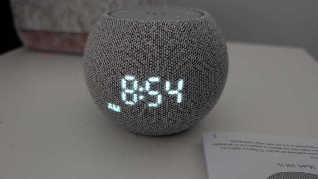 L'appareil est posé sur une table de chevet avec l'heure 8:54 affichée. Un manuel est visible en bas à droite.
